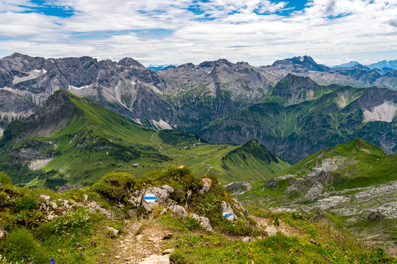 Ein schmaler Wanderweg schlängelt sich durch eine grüne Hügellandschaft mit wilden Blumen und Felsen.