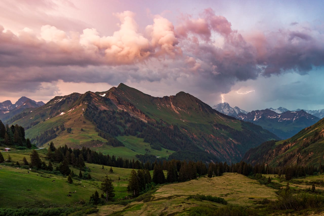 Eine Berglandschaft der Alpen mit grünen Tälern, schroffen Gipfeln und tiefhängenden Gewitterwolken, mit Blitzeinschlag.