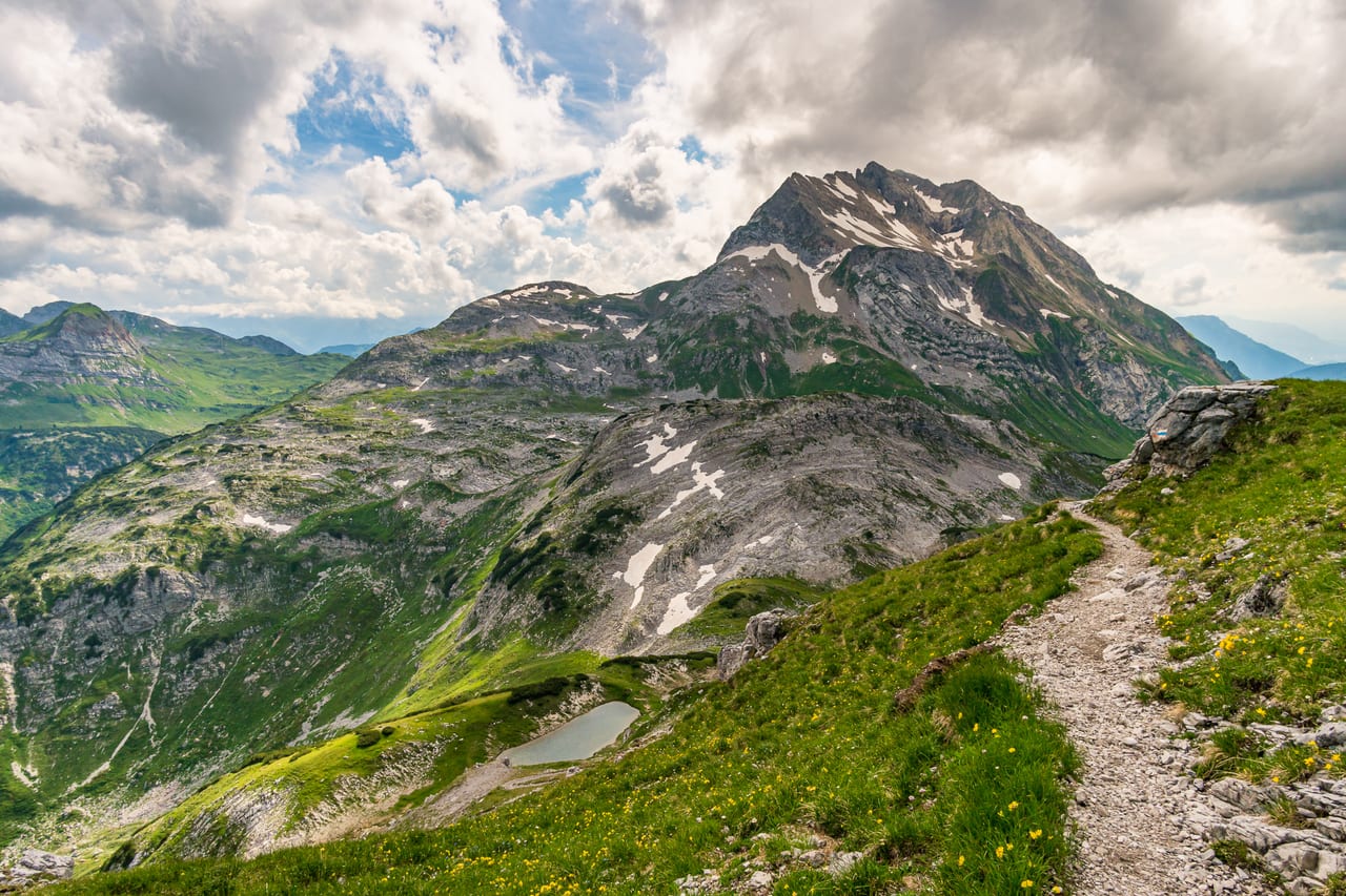 Ein malerischer Bergpfad schlängelt sich durch grüne Almen und steile Felsformationen.