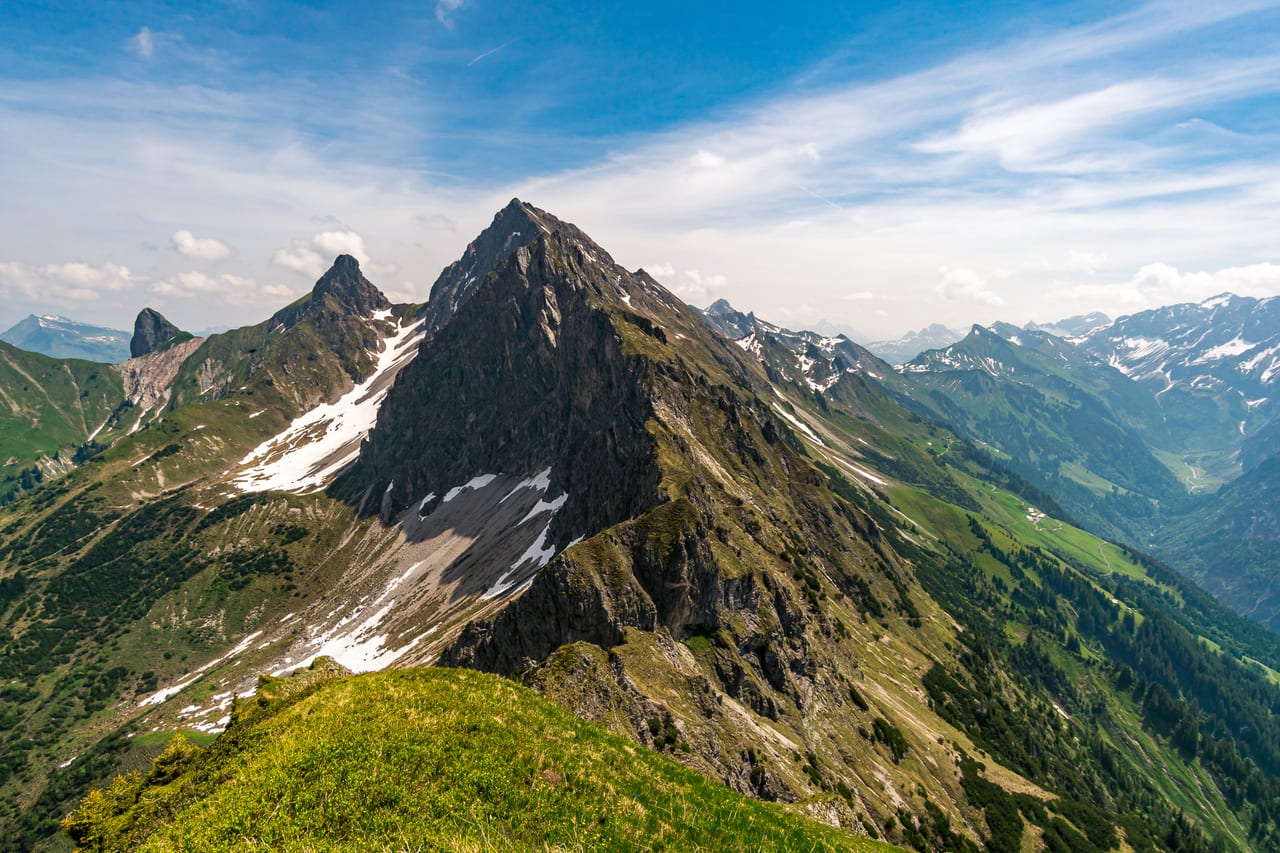 Ein majestätischer Berg mit schroffen Felsformationen und kleinen Schneeflecken ragt aus einem grünen Hochtal heraus.