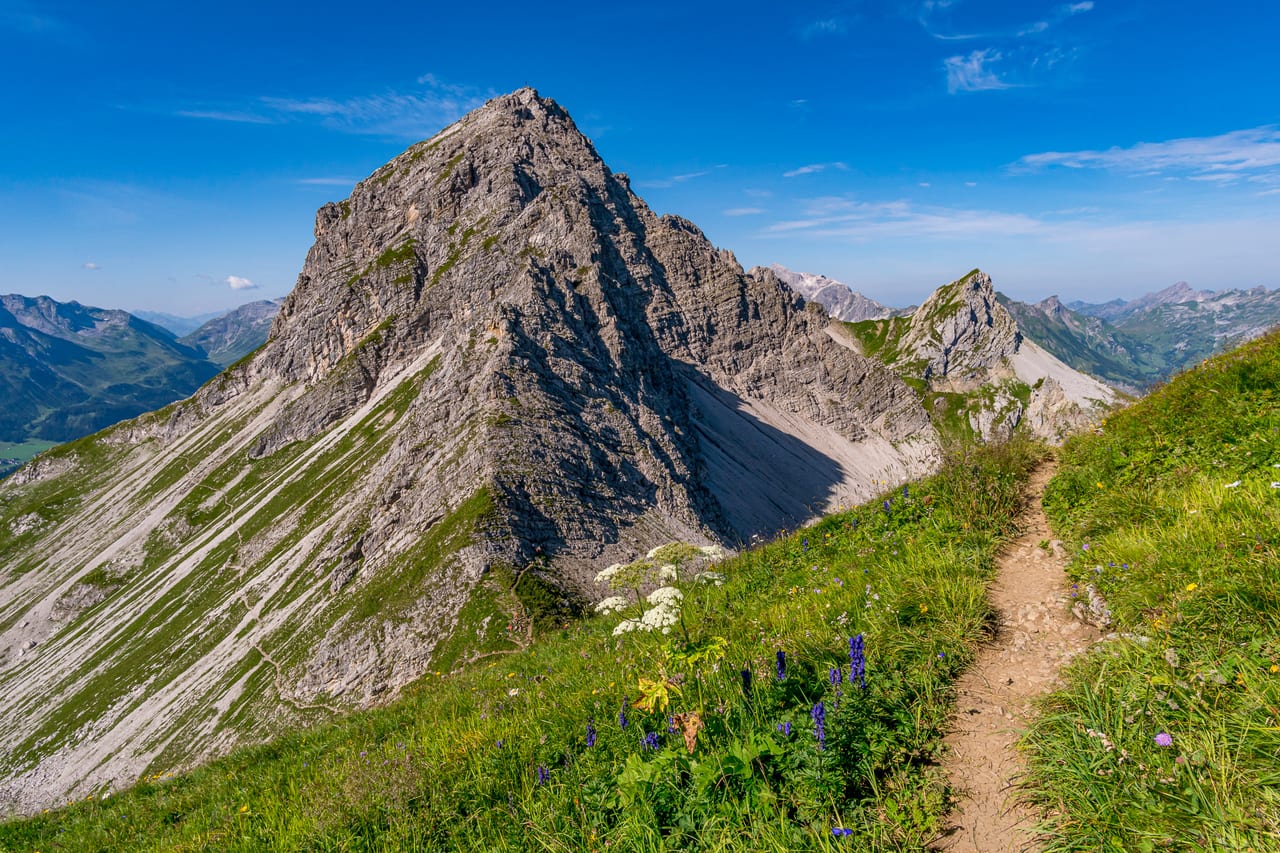 Ein steiler Berggrat der Alpen mit schroffen Felsformationen und einem schmalen, von wilden Blumen gesäumten Wanderweg.