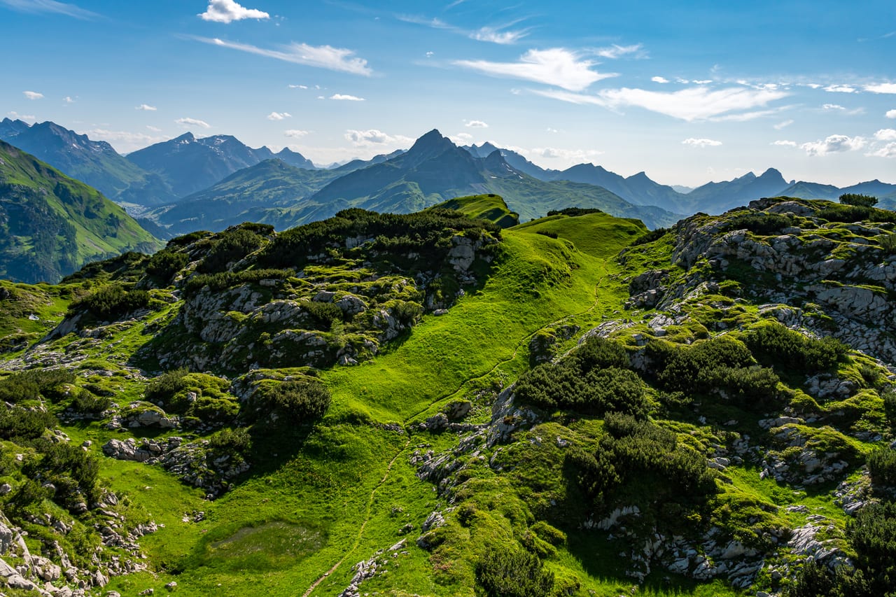 Eine weite, grüne Hochebene in den Alpen mit sanften Hügeln, Felsformationen und einem schmalen Pfad.