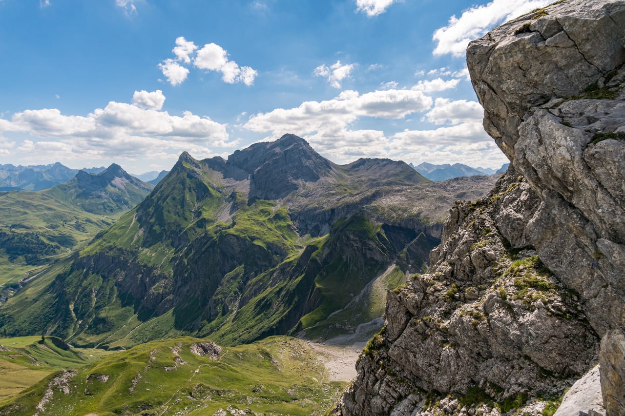 Ein weites Panorama der Alpen zeigt sich mit schroffen Felsvorsprüngen im Vordergrund, grüne Hänge.