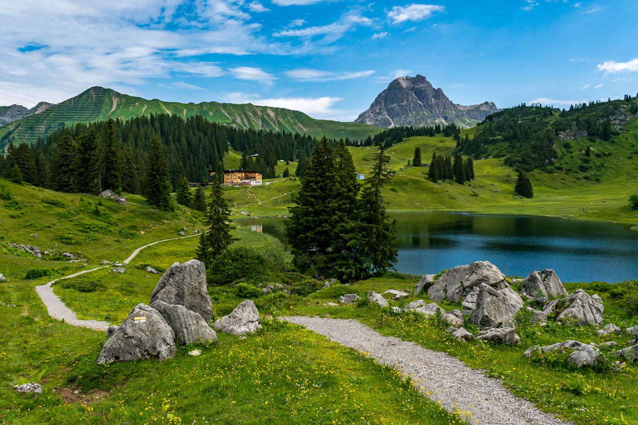 Ein klarer Bergsee in den Alpen, umgeben von grünen Wiesen, Fichtenwäldern und markanten Felsformationen.