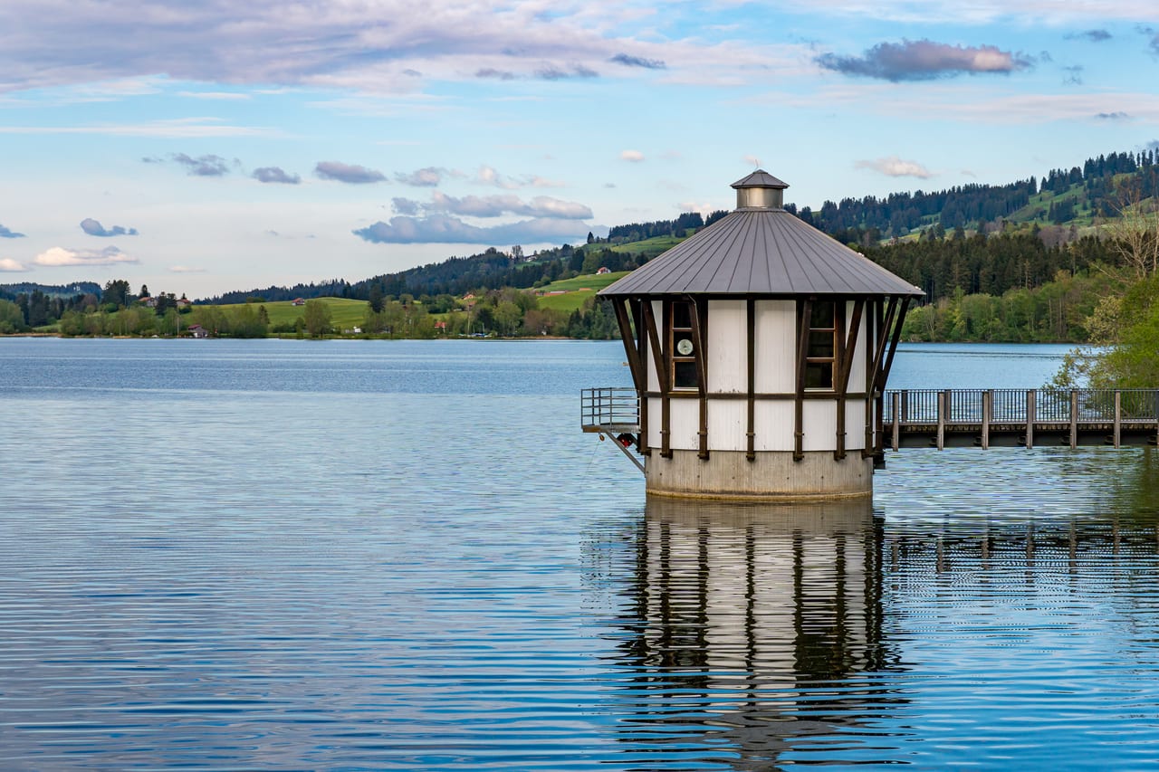 Ein kleines, oktogonales Bootshaus mit Metallspitze steht auf einer Plattform inmitten eines ruhigen Sees.