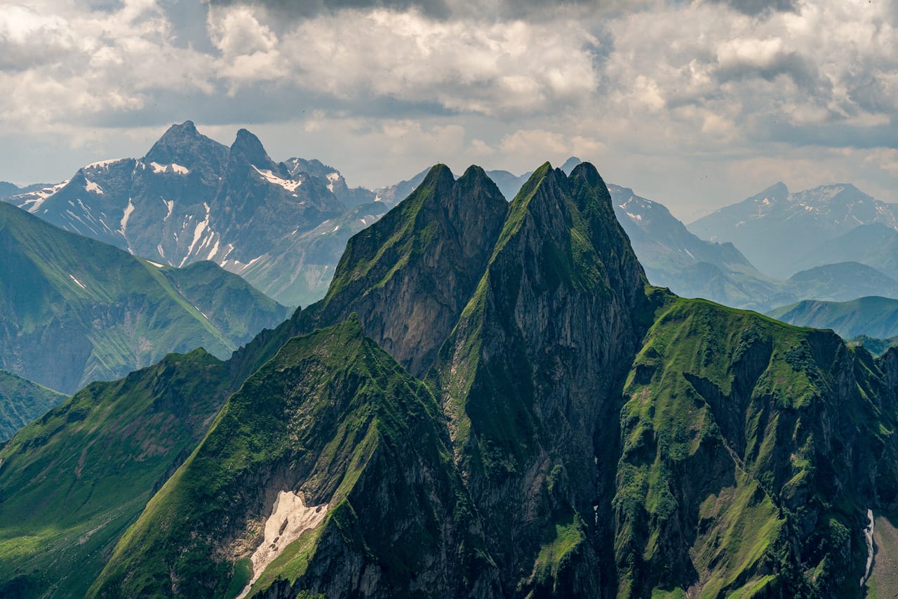 Drei markante, grüne Bergspitzen mit steilen Felsflanken ragen sich vor einem Hintergrund aus schneebedeckten Felsformationen.