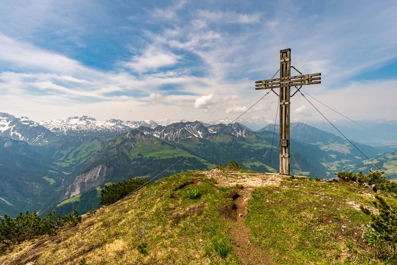 Ein großes Holzkreuz steht auf einem grasbewachsenen Bergplateau mit Blick auf eine weite, schneebedeckte Bergkette.