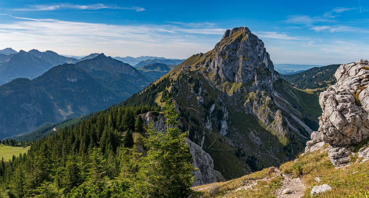 Ein markanter Felsgipfel ragt aus einem dichten Waldgebiet der Alpen empor, umgeben von schroffen Bergketten.