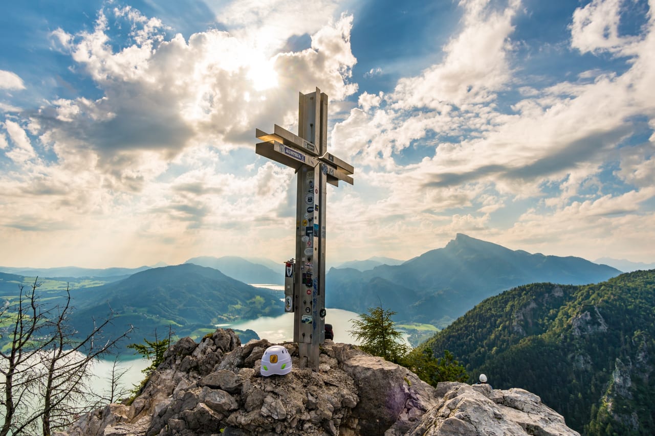 Ein großes Gipfelkreuz auf einem Felsen mit Blick auf ein tiefblaues Tal, umgeben von Bergen .