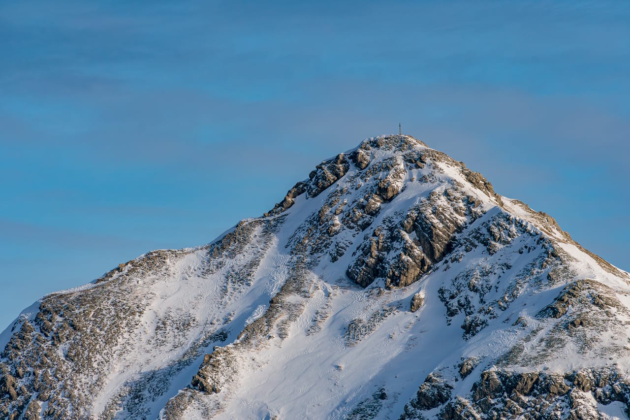 Eine schneebedeckte Bergspitze mit markanten Felsformationen und einem kleinen Gipfelkreuz auf dem höchsten Punkt.