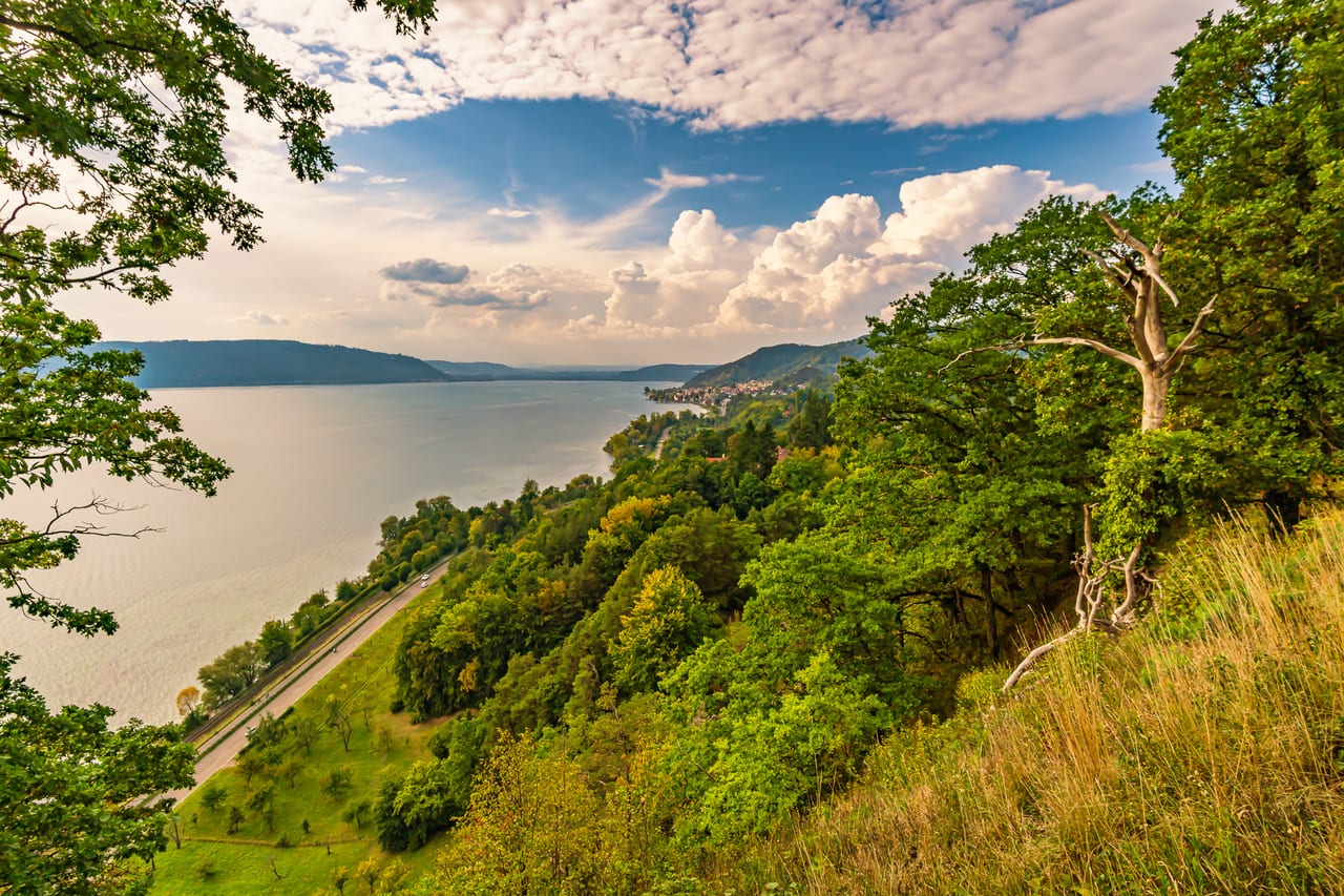 Eine malerische Landschaft mit einem ruhigen See, der sich zwischen grünen Hügeln und bewaldeten Berghängen erstreckt.