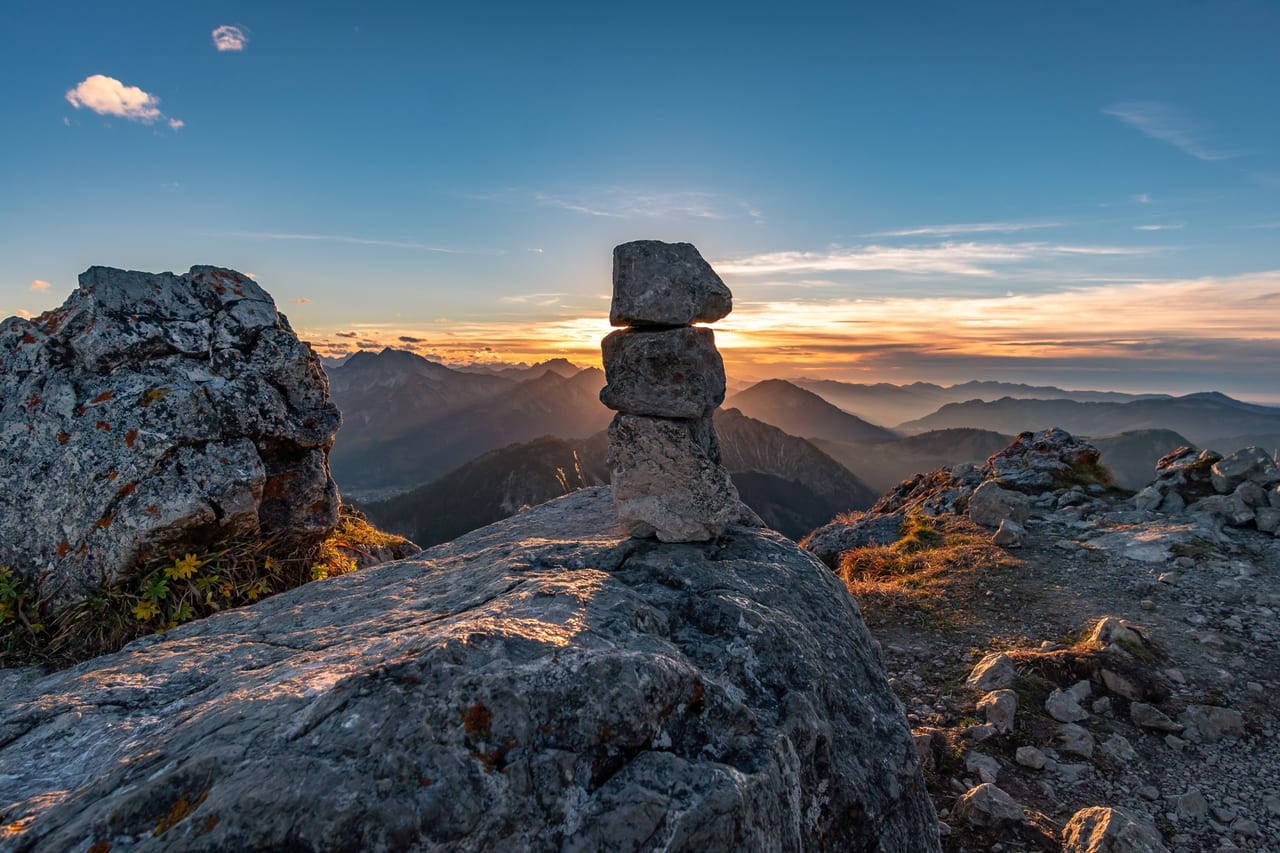 Eine sorgfältig gestapeltes Steinmännchen auf einem felsigen Gipfel, im Hintergrund eine weite Berglandschaft.