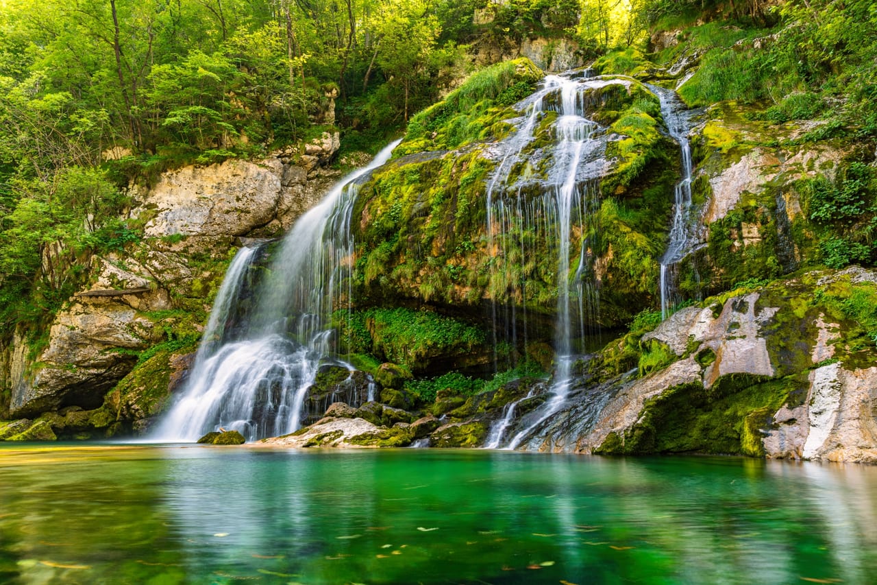 Kräftiger Wasserfall stürzt sich über moosbewachsene Felsformationen in einen klaren, smaragdgrünen Bergbach.