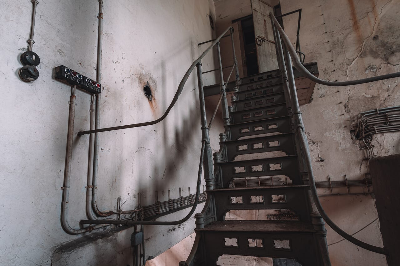 Eine abgenutzte Metalltreppe in einem verlassenen Industriegebäude, umgeben von rostigen Rohrleitungen.