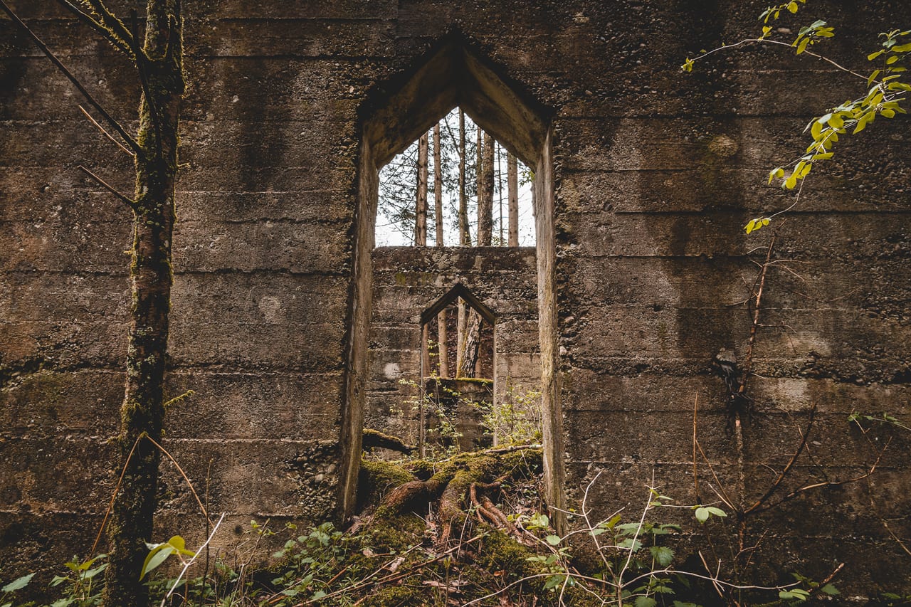 Eine verwitterte Steinmauer mit einem halbzerstörten, gotisch anmutenden Fenster, das von Moos und Pflanzen überwuchert.