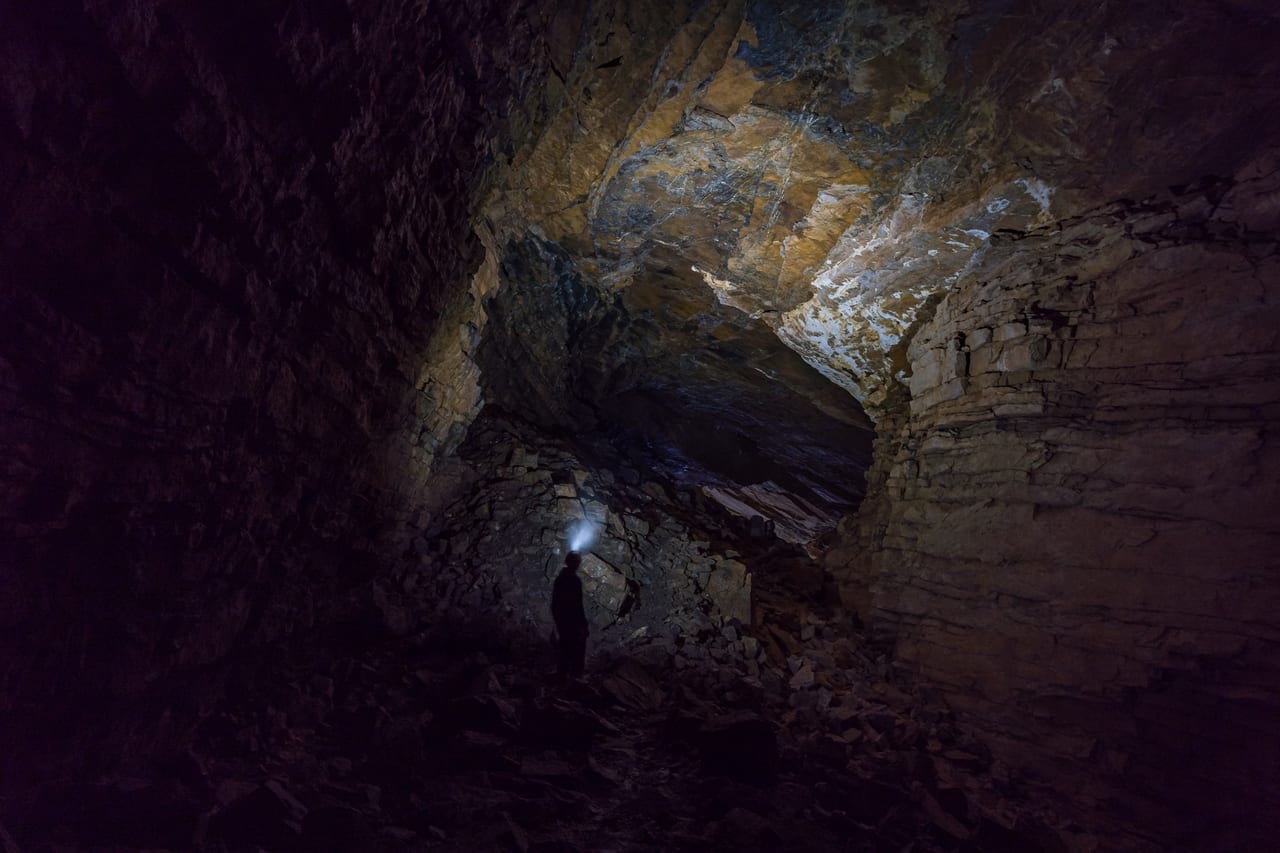 Eine Person mit Kopflampe erkundet einen dunklen, verlassenen Bergschacht mit moosbewachsenen Wänden.