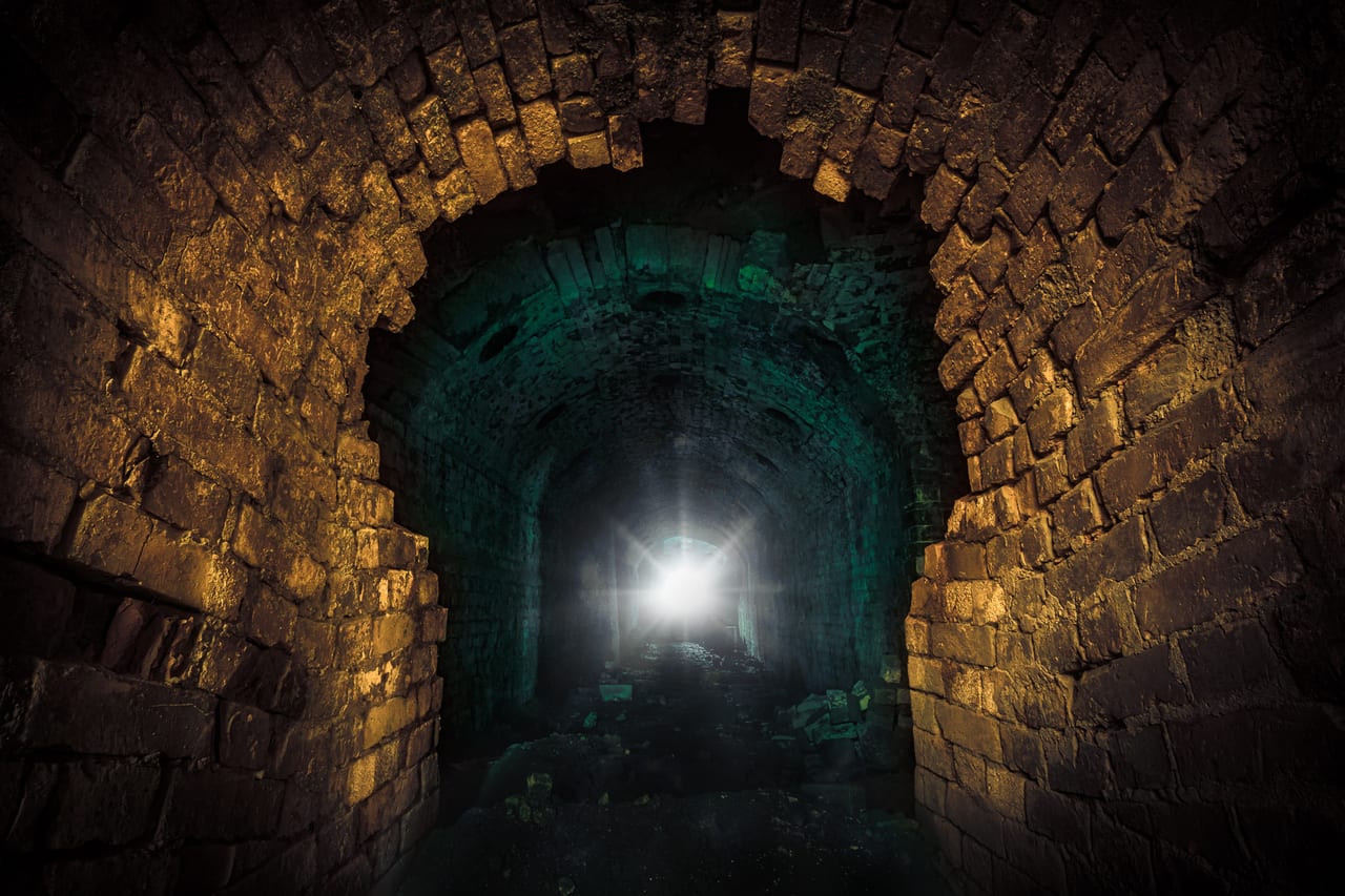 Ein alter, aus groben Natursteinen gemauerter Tunnel fährt ins Dunkel, mit einer fernen Lichtquelle am Ende des Gangs.