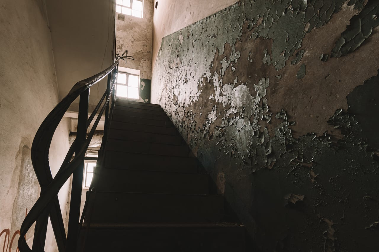 Eine abgenutzte, schmale Treppe führt in einem verfallenen Gebäude mit stark beschädigten, moosbewachsenen Wänden.