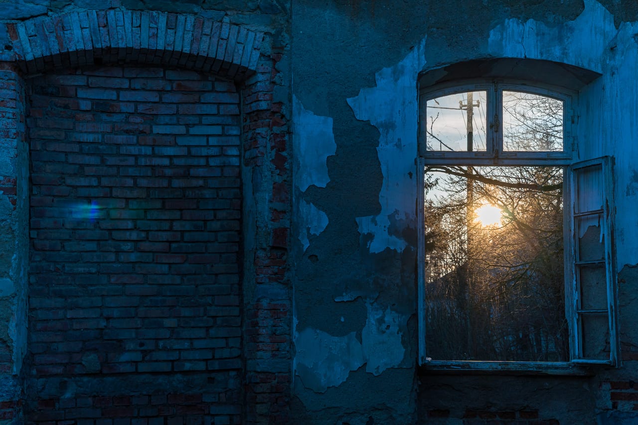Ein verfallenes Bahnhofsgebäude mit abblätternder blauer Wandfarbe und zwei offenen Fenstern, durch die die untergehende Sonne strahlt.