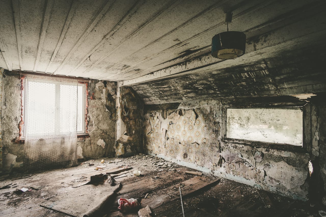 Ein verlassener Raum mit stark beschädigten Wänden, abblätternder Tapete und zerbrochenen Fliesen.