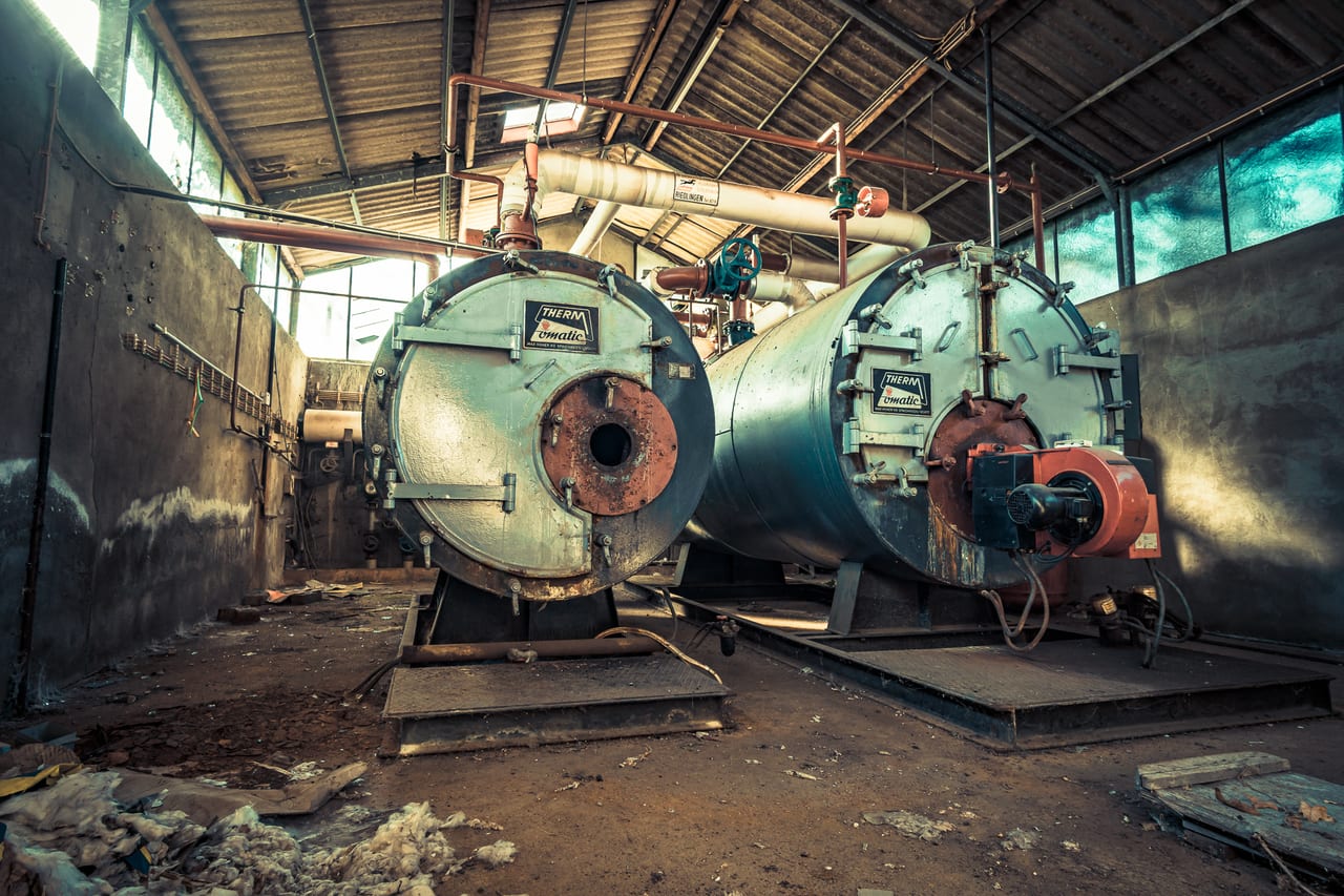 Zwei große, rostige Dampfkessel stehen in einer verlassenen und verfallenen Industriehalle.