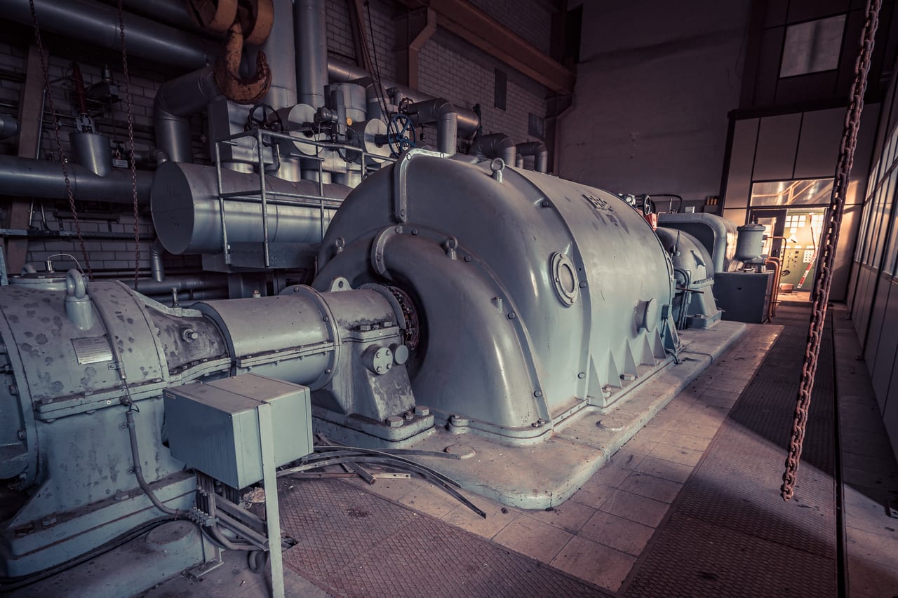 Eine rostige und veraltete Dampfmaschine steht in einer verlassenen Industriehalle mit sichtbaren Rohren und Pipelines.
