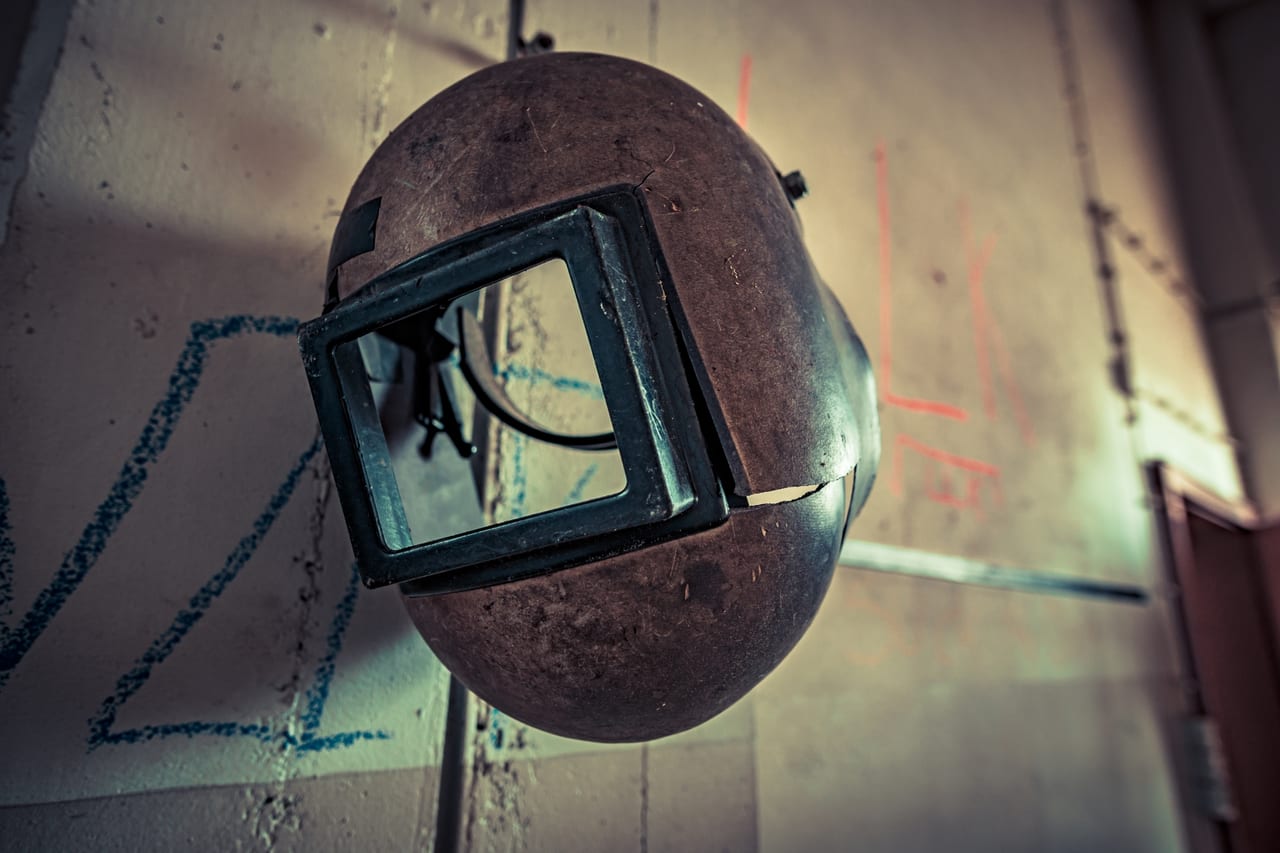 Ein stark korrodierter, runder Schweißerhelm mit quadratischem Glasfenster hängt schräg von einer Decke in einer.