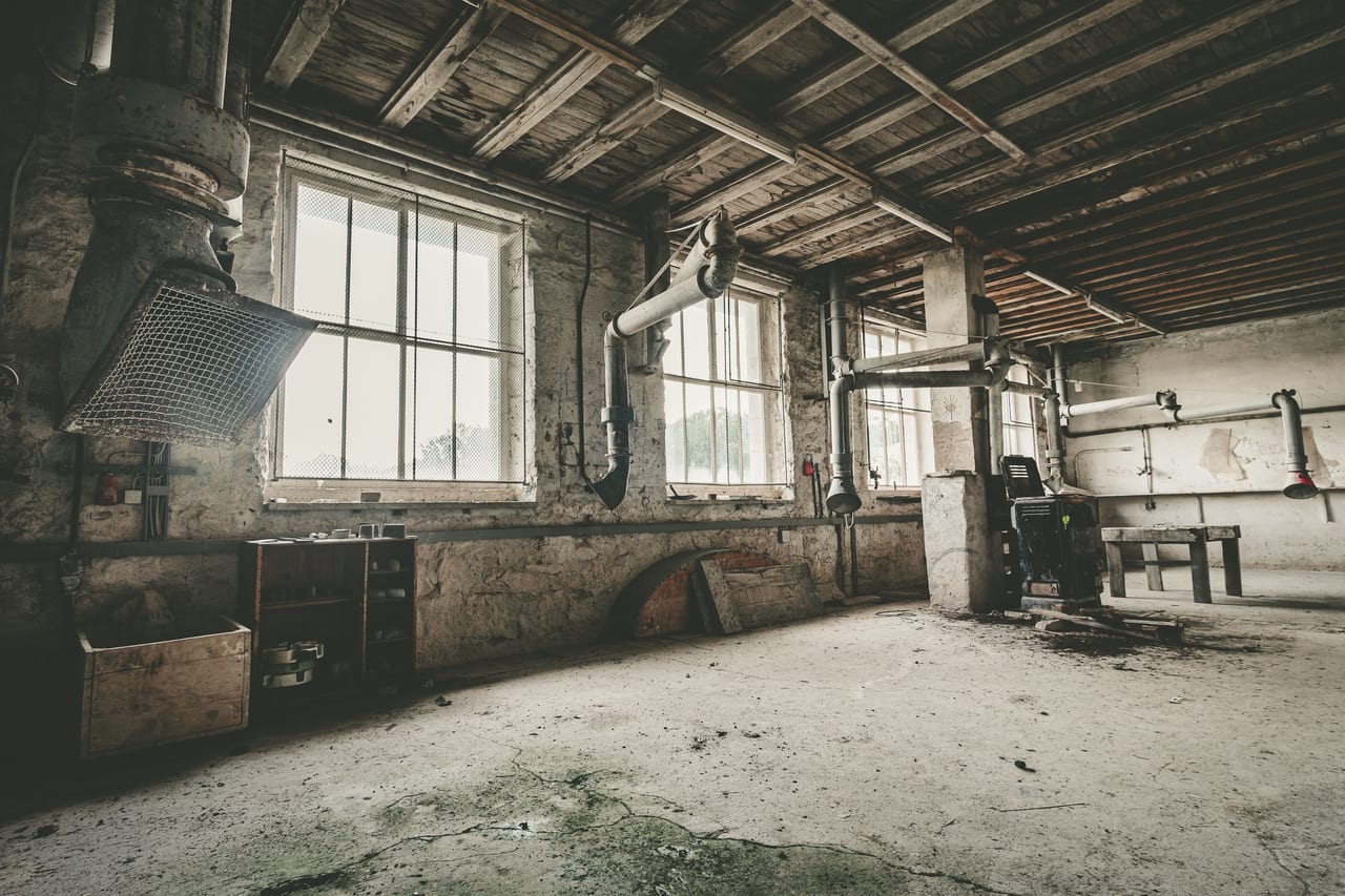 Eine verlassene, leere Steinmetz Werkstatt mit Betonpfeilern, alten Metallrohren und großen Fenstern.