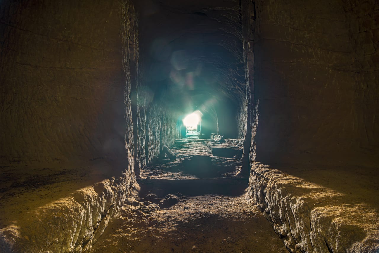 Ein verlassener, dunkler Tunnel aus Beton und Stein mit einer hellen Lichtquelle am Ende, die durch eine Öffnung scheint.