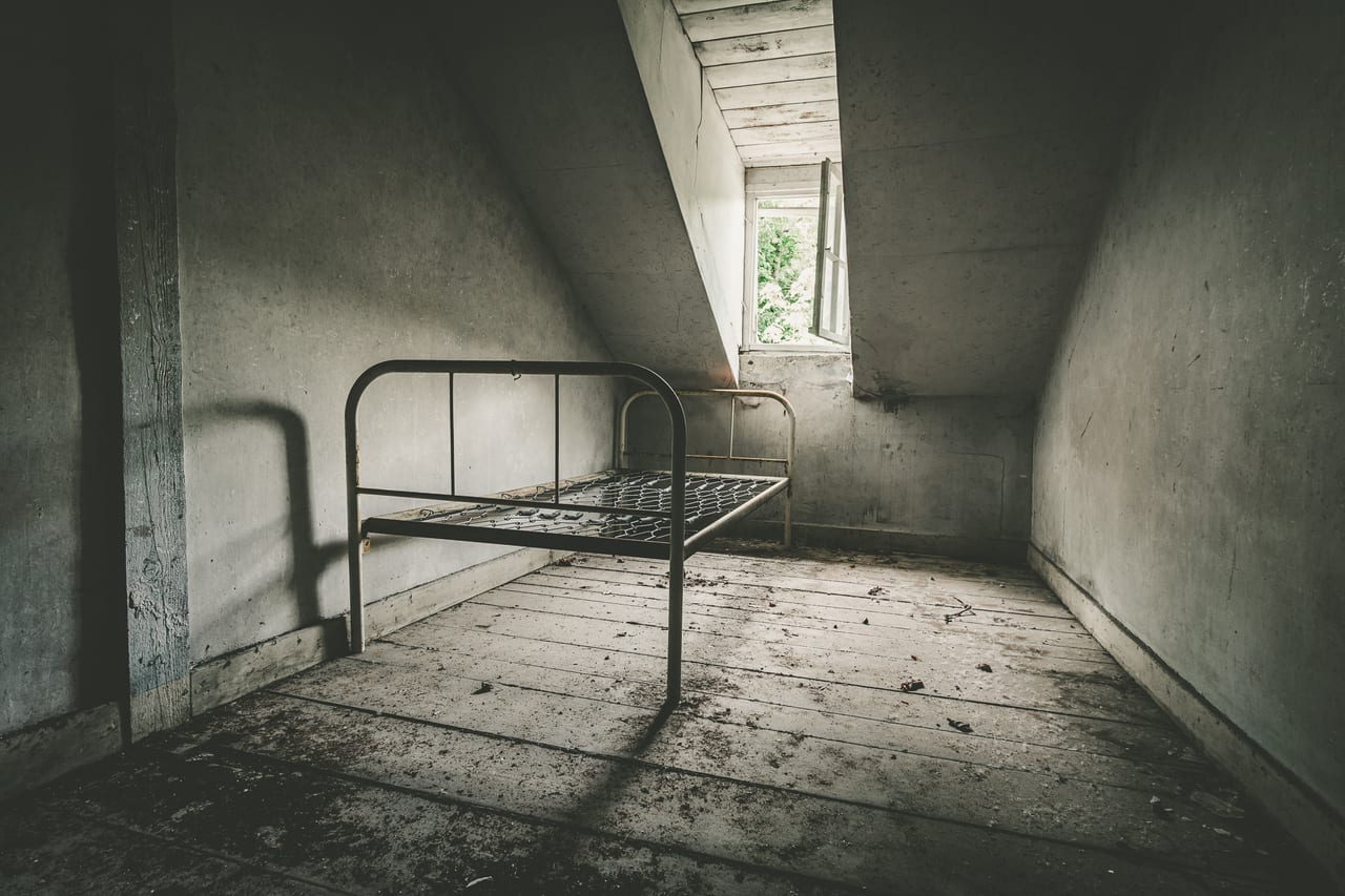 Ein verlassener Raum unter einem schrägen Dach mit abblätternder Wandfarbe, einem rostigen Metallbett.