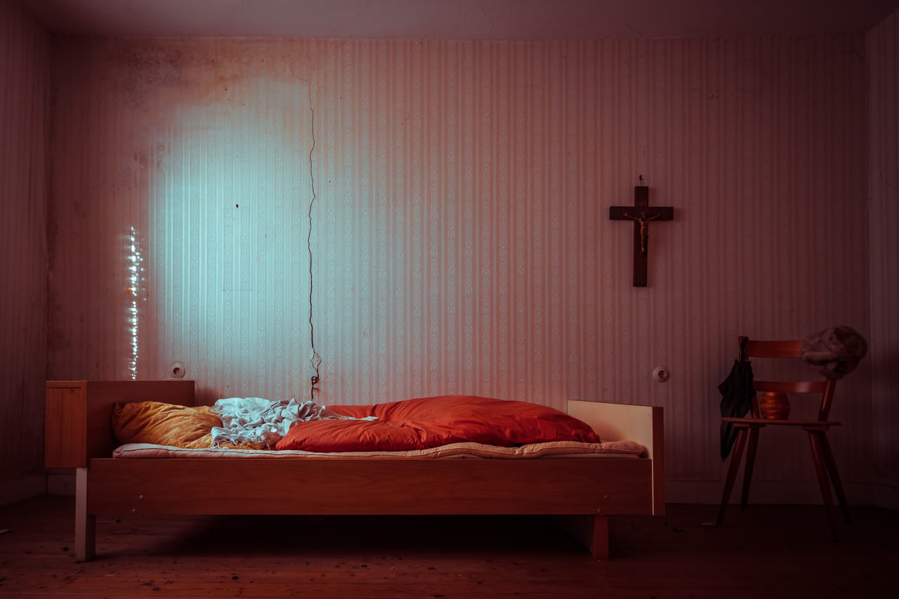 Ein verlassener Raum mit abblätternder Wandtapete, einem ungemachten Bett mit orangefarbenem überzug und einem Kreuz an der Wand.